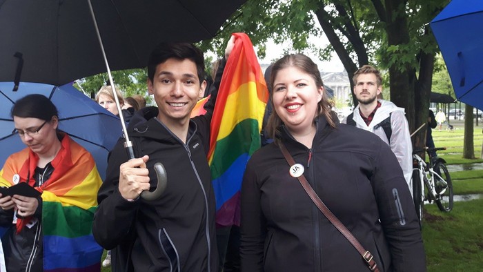 Eine Gruppe von Menschen steht im Regen, einer hält einen Regenbogen-Schirm.