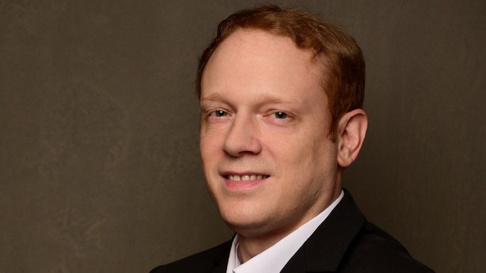 Johannes Bernhard Bernarding in einem formellen schwarzen Anzug und weißem Hemd posiert selbstbewusst mit verschränkten Armen.