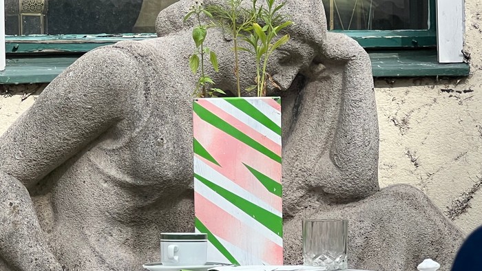 Eine Steinstatue eines Mannes, der sich auf seinen Ellbogen stützt, mit einer gestreiften Geschenkbox und einer Pflanze auf seinem Schoß.