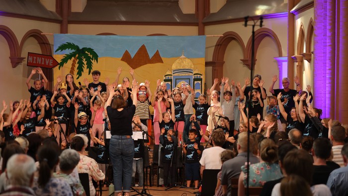 Eine lebhafte Gruppenaufführung mit Kindern und Erwachsenen, die begeistert auf einer geschmückten Bühne singen.