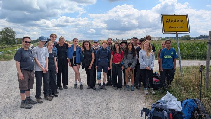 Gruppe von Menschen, die an einem sonnigen Tag im Freien in der Nähe eines Straßenschilds mit der Aufschrift „Altötting“ posieren.
