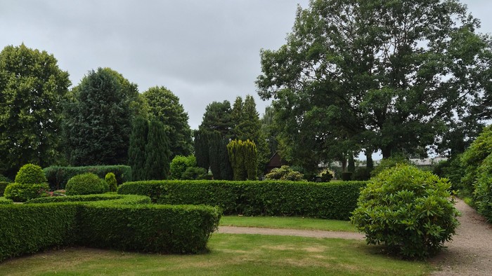 Üppiger Gartenweg, gesäumt von akkurat geschnittenen Hecken und grünen Sträuchern unter bedecktem Himmel.