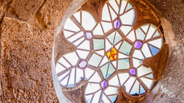 Ein ornamentales Kirchenfenster mit kleinen farbigen Glasflächen ist in eine dicke steinerne Fensterlaibung eingelassen. Das einfallende Tageslicht hebt die geometrischen Formen und die Struktur des Mauerwerks hervor.