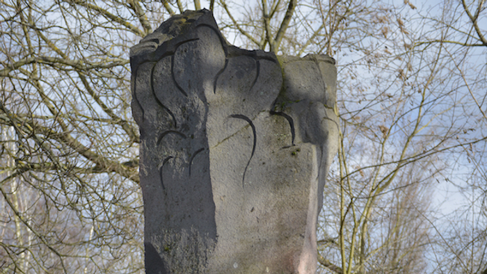 Steinsäule mit aufwendigen Schnitzereien vor einem Baumhintergrund.