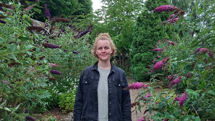 En person står og smiler på en grussti omgivet af livligt lilla blomstrende planter.