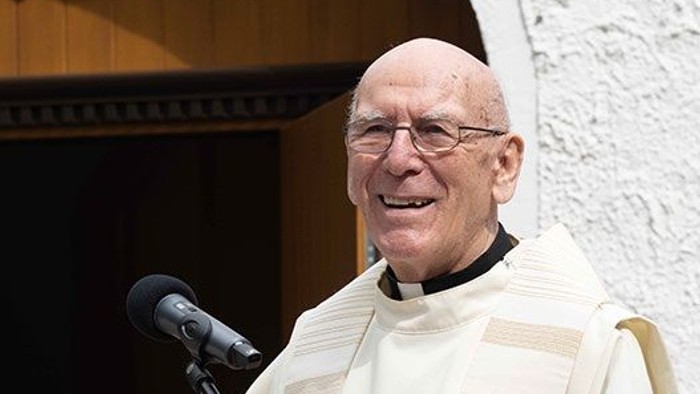 Ein kahlköpfiger Mann in geistlichem Gewand lächelt, während er in ein Mikrofon spricht.