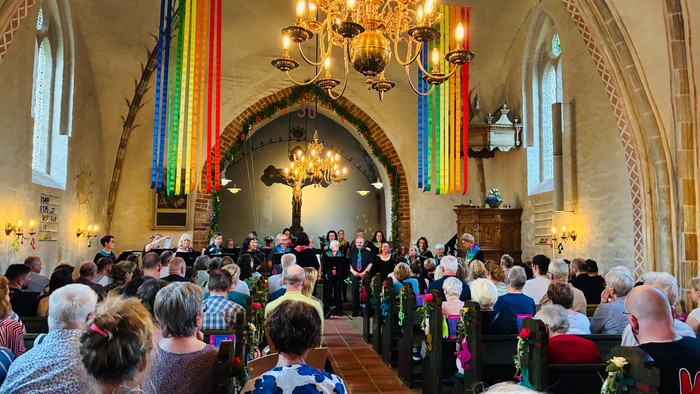 Eine vielfältige Menschenmenge versammelt sich in einer historischen Kirche zu einer Choraufführung mit gezeigten Regenbogenfahnen.