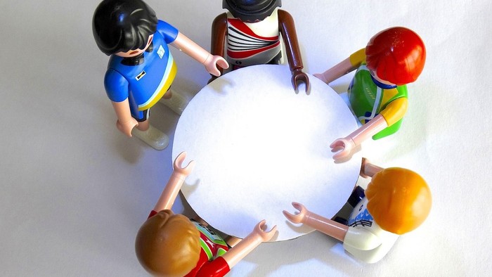 Der Betrachter blickt von oben auf fünf Playmobil-Figuren, die um einen runden Tisch versammelt sind.