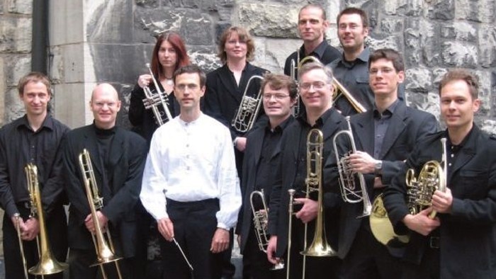 Ein Blechbläserensemble von Musiker:innen, das sich im Freien mit ihren Instrumenten zusammen aufstellt.