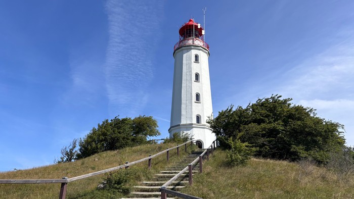 Ein malerischer weißer Leuchtturm steht auf einem grasbewachsenen Hügel, zu dem eine Treppe hinaufführt.