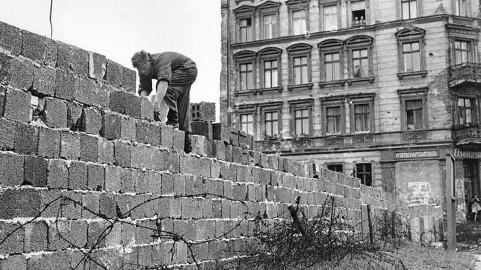 Eine Person klettert an einer mit Stacheldraht gesicherten Betonmauer neben einem alten mehrstöckigen Gebäude.