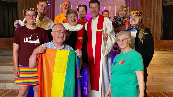 Bunt angezogene Menschen, ausgestattet mit Fahnen in Regenbogenfarben, stehen in der St.-Marien-Kirche und lachen in die Kamera.
