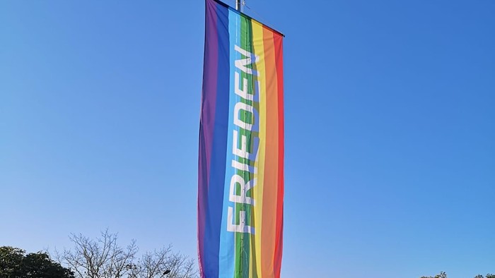 Regenbogenflagge mit der Aufschrift „FRIEDEN“ (Deutsch für Frieden), die vor einem klaren blauen Himmel weht.