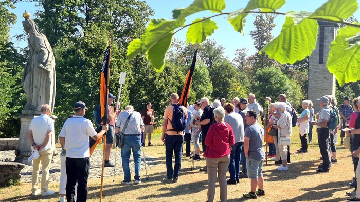 Gruppe von Menschen, die sich im Freien in der Nähe einer Statue versammelt haben und in einer sonnigen Parklandschaft Flaggen halten.