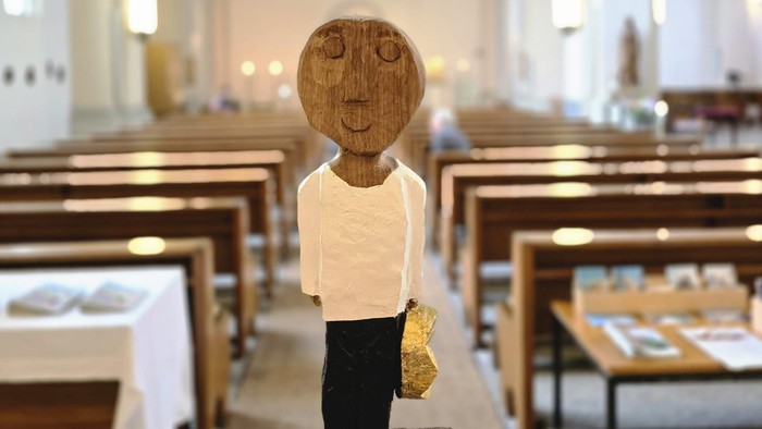 Holzfigurine im Kirchenschiff, die einen kleinen Gegenstand hält, auf einem Steinsockel platziert.