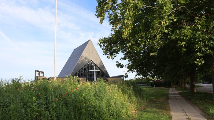 En moderne kirke med trætag og et kors på fasaden, omgivet af græs og træer