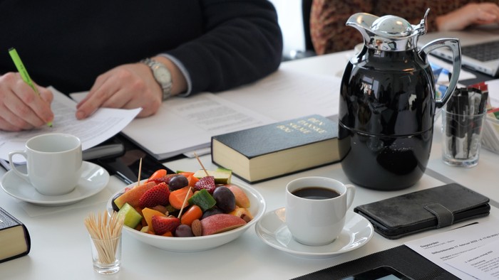 To dagslys og en gruppe af mennesker sidder ved et bord med notater, en kaffe-thermos, en fruktsalat og andre drikkevarer.