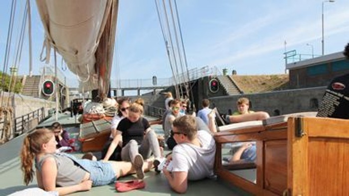 Gruppe entspannt auf dem Deck eines hölzernen Segelboots in der Nähe einer malerischen Uferpromenade mit historischen Gebäuden im Hintergrund.