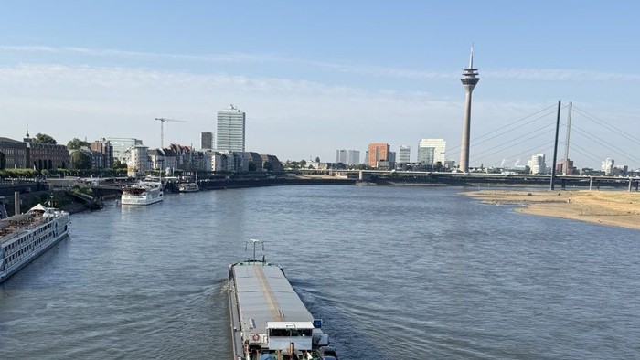Ein Boot fährt einen Fluss entlang mit einer Stadtkulisse und einem hohen Turm im Hintergrund unter klarem Himmel.