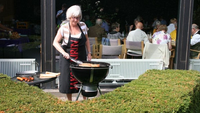 En kvinde ståer ved grill i trætern med rødt og svart mønster på skjorte og sorte bukser.