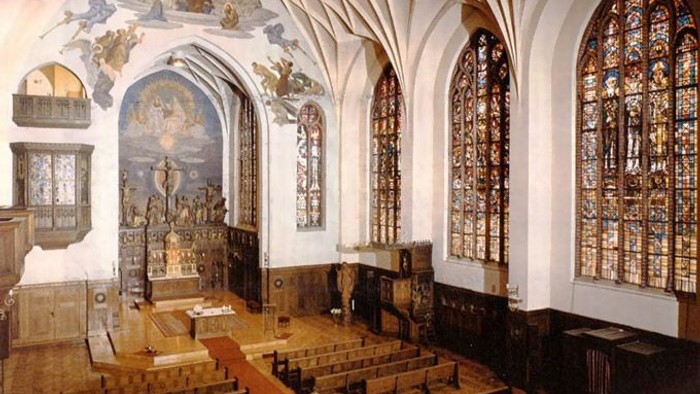 Historische Kapelle mit kunstvollen Buntglasfenstern, hölzernen Kirchenbänken und aufwendigen religiösen Fresken an gewölbten Decken.