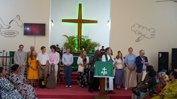 Gruppe von Jugendlichen und Erwachsenen im Altarraum einer Kirche in Tanga. Im Hintergrund befindet sich ein großes beleuchtetes Holzkreuz, während ein Mann mit Mikrofon zur Gemeinde spricht.