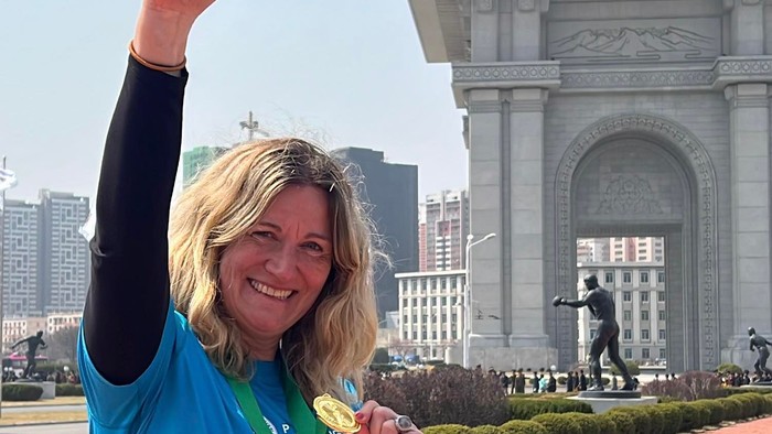 Glædestrålende løber viser stolt medalje og startnummer frem foran et stort monument.