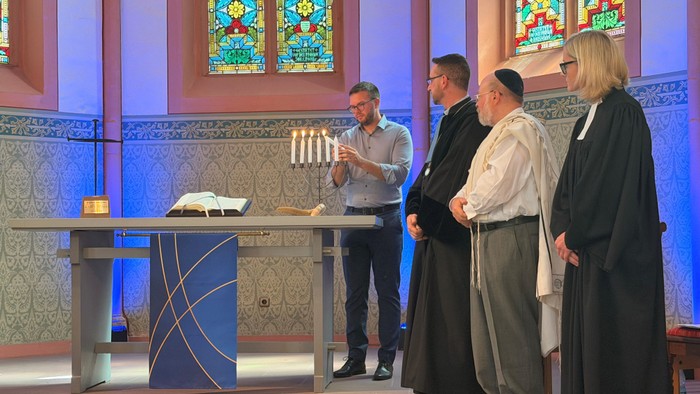 Ein Mann entzündet in einer blau beleuchteten Kirche eine Kerze an einer Menora, während neben ihm ein evangelischer Geistlicher, ein jüdischer Geistlicher mit Kippa und Gebetsschal sowie eine evangelische Pfarrerin vor den farbigen Kirchenfenstern stehen
