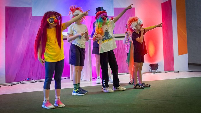 Vier kostümierte Kinder vor einer bunten Wand zeigen in die Höhe