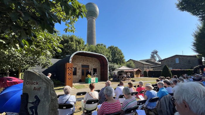 Hier sieht man das Gelände vom Knappenverein mit dem Übungsstollen und einem Wasserturm im Hintergrund, Vorne sitzen Besucher des Gottesdienstes.