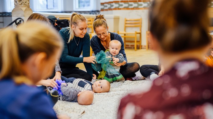 En gruppe kvinder sidder på gulvmatte og omhandler børnepuppetter.