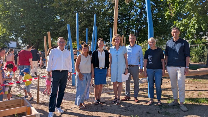 Auf dem Außengelände einer Kita stehen sieben Erwachsene nebeneinander vor einem neuen Kletter- und Spielbereich mit hohen blauen und naturfarbenen Holzstämmen. Im Hintergrund spielen zahlreiche Kinder und Erwachsene auf dem Gelände.