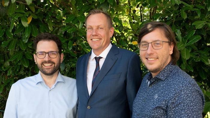 Drei Männer mittleren Alters stehen in Hemd und Anzug vor einer grünen Hecke und lächeln.