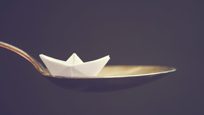 Ein Papierboot balanciert zart auf einem Löffel vor dunklem Hintergrund.