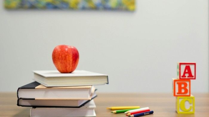 Eine rote Apfel liegt auf gestapelten Büchern neben bunten Buchstabenbausteinen und Bleistiften auf einem Schreibtisch.