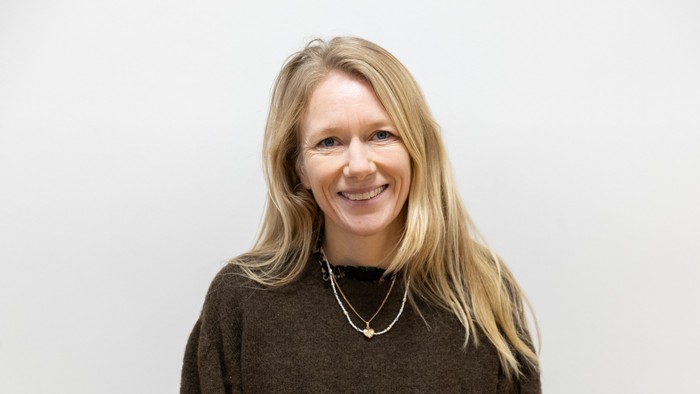 En glad kvinde med blondt hår iført en brun sweater og et halskæde smiler til kameraet.