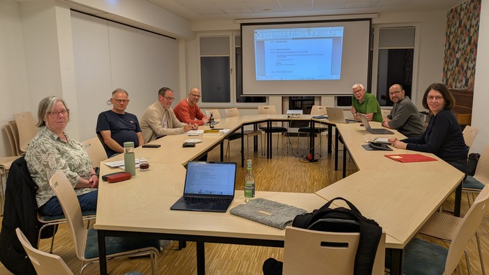 Eine Gruppe von Personen versammelte sich um einen Konferenztisch in einem Besprechungsraum mit einer Präsentation auf dem Bildschirm.