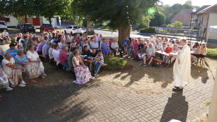 Eine große Outdoor-Veranstaltung mit Menschen, die auf Bänken sitzen, während ein Priester in einer sonnigen Umgebung eine Zeremonie leitet.