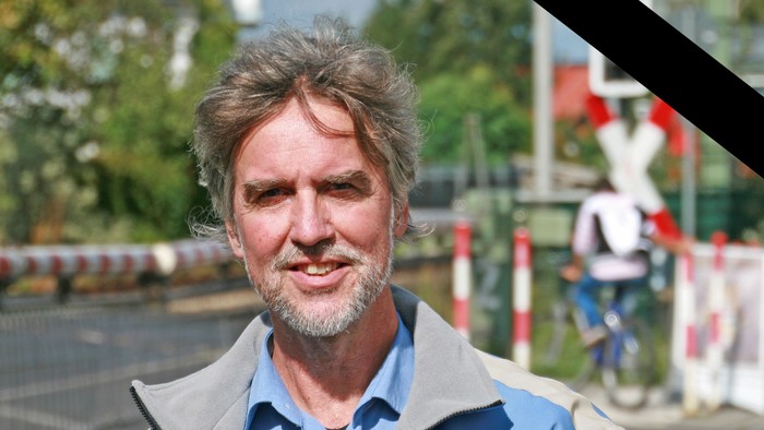 Ein Porträt eines grauhaarigen, bärtigen Mannes im Freien; im Hintergrund sind unscharf ein Bahnübergang und eine Person auf einem Fahrrad zu sehen. Ein schwarzes Trauerband verläuft diagonal über die rechte obere Bildecke.