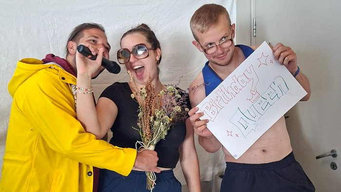 Drei Personen feiern einen Geburtstag mit einem freizügigen Schild, getrockneten Blumen und verspieltem Gesichtsausdruck.