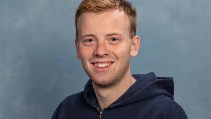 Junger Mann, der in einem marineblauen Hoodie vor einem blauen Hintergrund lächelt.