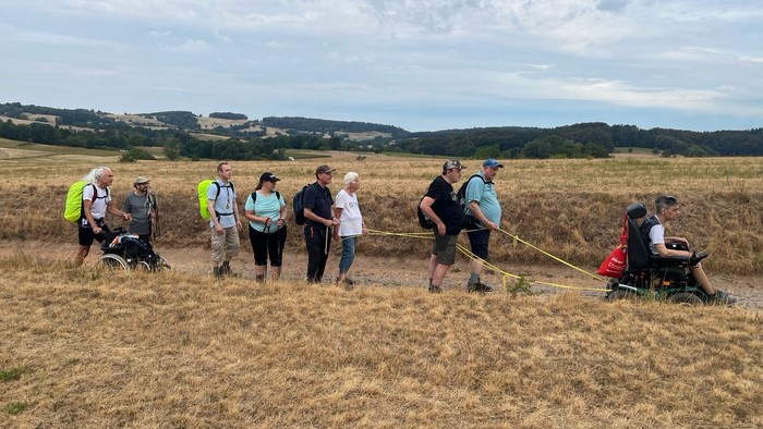 Eine Gruppe Pilgernde zieht in einer Reihe über einen staubigen Weg durch hellgelbe Feldern. In der Ferne sind Hügel mit Wäldern zu sehen. Die Pilgergruppe ist leicht gekleidet, sie tragen Rucksäcke, sie sind bunt gekleidet. Vorne ein Elektrorollstuhlfahr