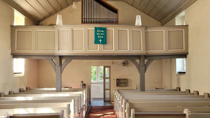 Ein ruhiger Kircheninnenraum mit hölzernen Kirchenbänken, einem roten Teppichgang und einer Orgel im erhöhten Altarraum.
