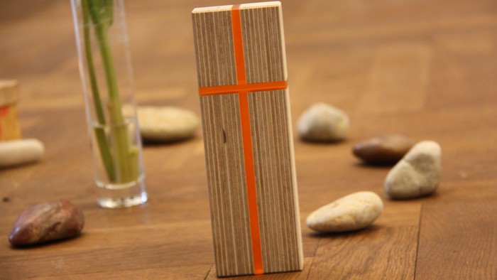Holzblock mit Kreuz auf Holzboden, dahinter Steine