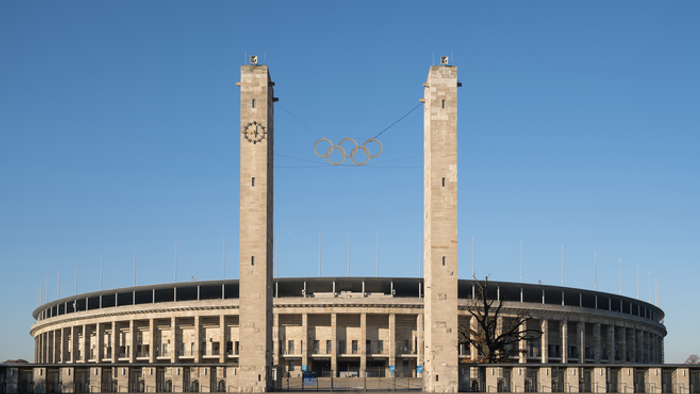 Haupteingang des Olympiastdions in der Totalen mit den den zwei herausragenden Türmen und dem gesamten Gebäude vor blauem Himmel