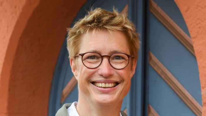 Eine fröhliche Person mit Brille, einem grünen Mantel und einem weißen Oberteil vor farbenfrohen architektonischen Bögen.