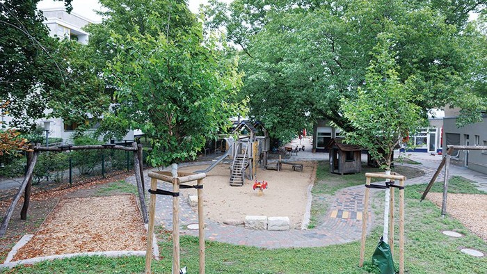 Ein schattiger Spielplatz mit Klettergerüsten, Rutschen und Sandbereich, umgeben von Bäumen und Bänken.