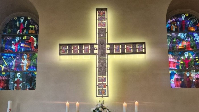 Eleganter Kirchenaltar mit Buntglasfenstern, Kerzen und einem zentralen Kreuz, das von oben beleuchtet wird.