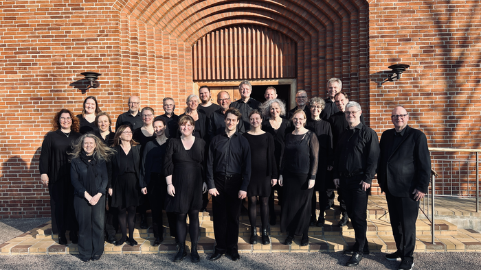 En gruppe musikere i formelt sort tøj poser sammen foran indgangen til en murstensbygning.