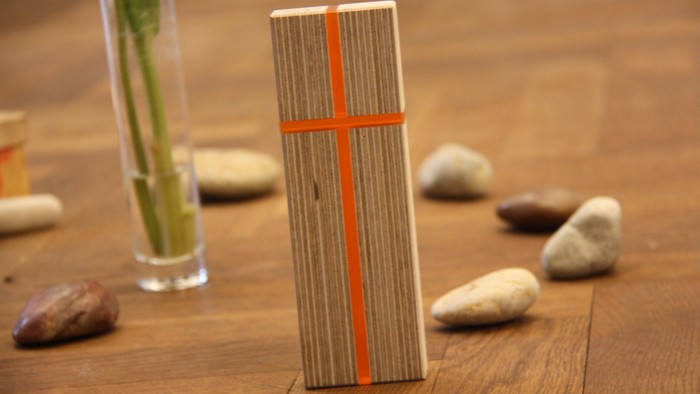 Kreuz steht schräg auf dem Boden, darum herum einzelne Steine und eine Vase mit Blütenstengeln.
